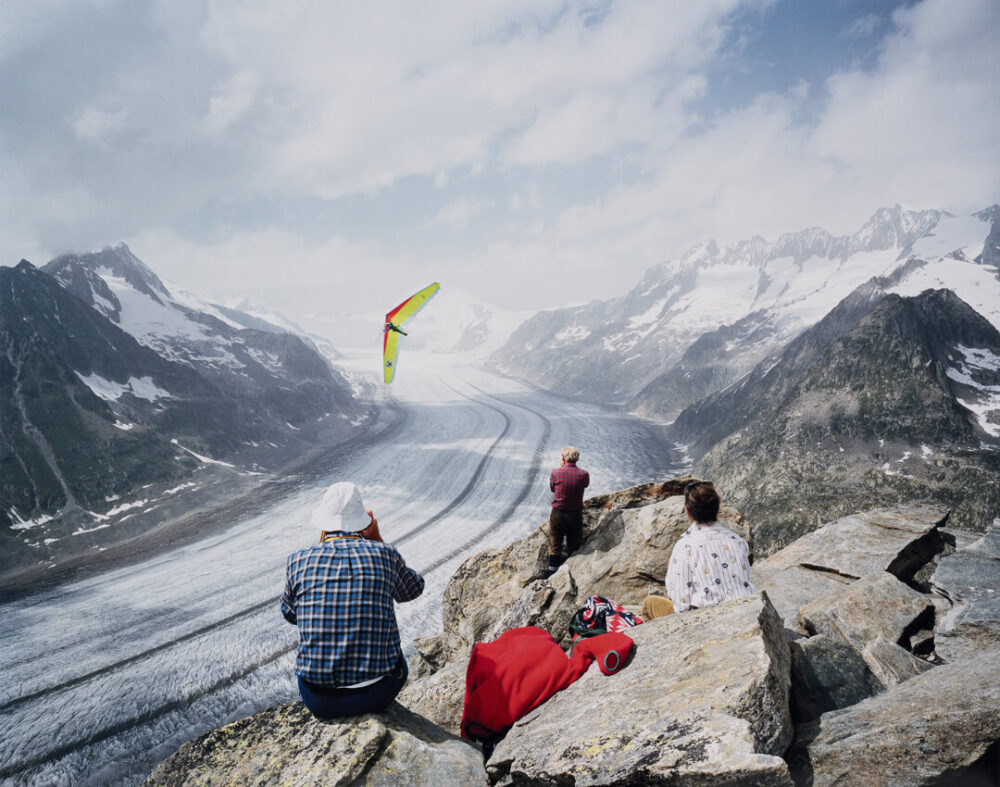 Fotografie des Alteschgeltschers mit drei Wander:innen und eine Hängegleiter fliegenden Person in der Luft von Nicolas Faure mit dem Titel 