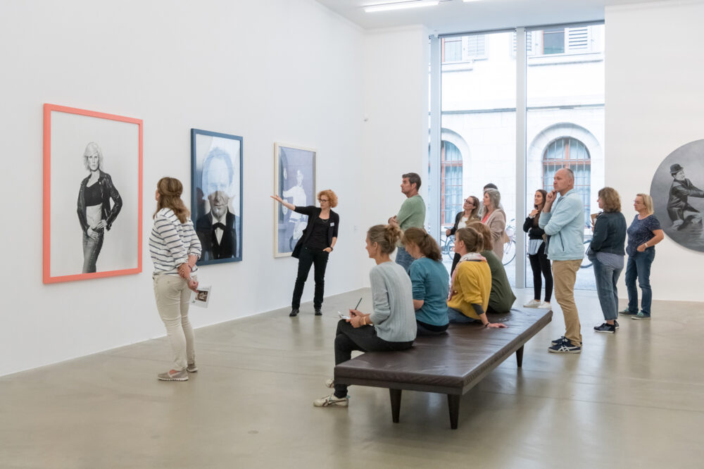 Eine Gruppe von Lehrpersonen steht in einer Fotoausstellung und wird von einer Vermittlungsperson eingeführt