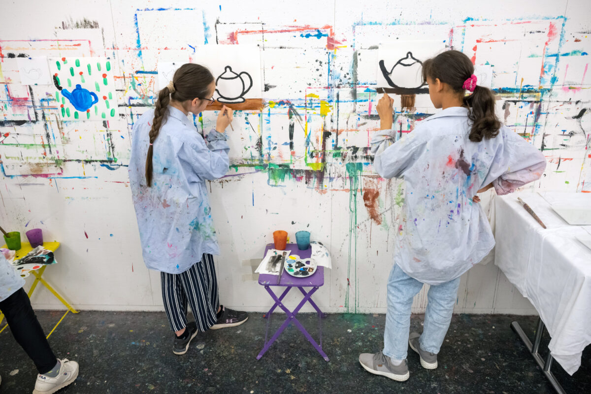 Zwei Kinder, die im Atelier an einer Wand etwas mit Pinsel malen