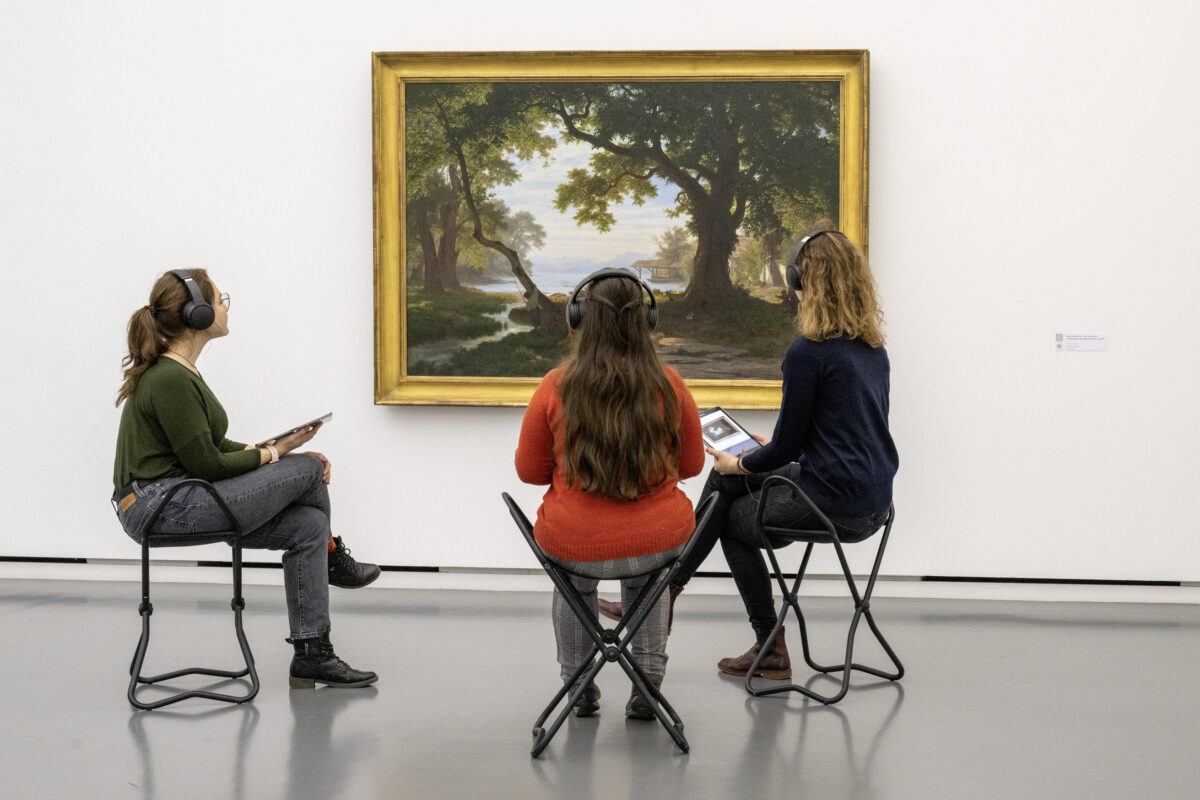 Drei Besucherinnen hören mit Kopfhörer eine Meditationsanleitung vor einem Gemälde