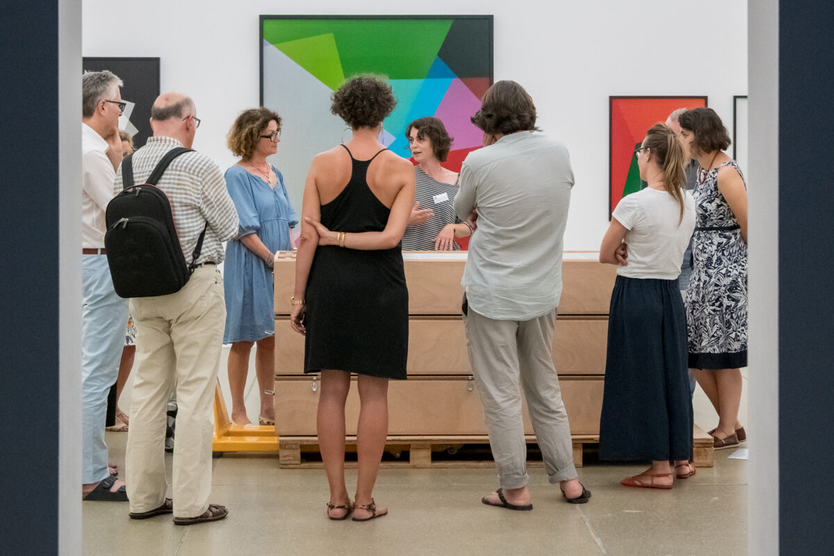 Personen stehen um eine Kiste mit Bildern, eine Restauratorin erklärt ihnen ihre Arbeit