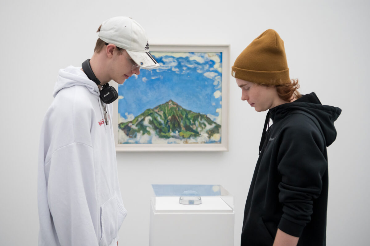 Zwei Schüler schauen ein Kunstobjekt in einer Vitrine an, dahinter ist ein Gemälde des Niesen (Berg in Thun) von Hodler zu sehen