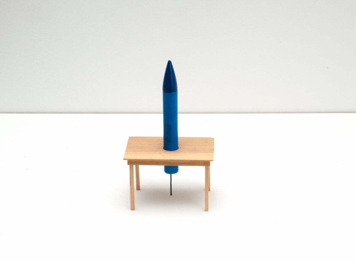 Skulptur einer Feuerwerksrakete, die in einem Tisch steckt.