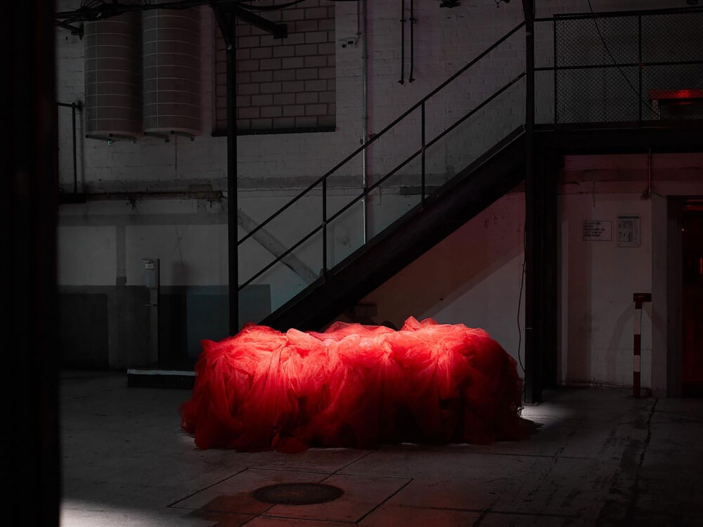 Foto eines länglichen Objektes, das unter einem roten Vorhang liegt und leuchtet in einer dunklen Industriehalle.