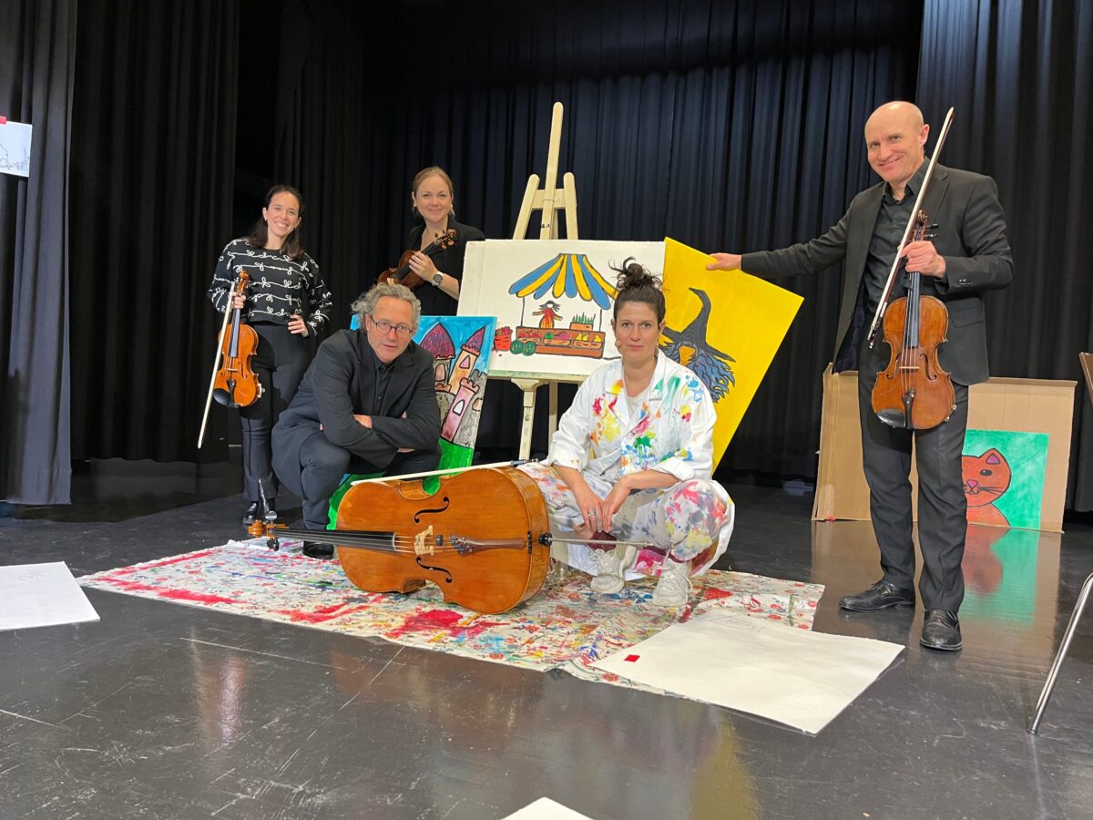 Eine Gruppe von Musikerinnen und Musikern mit Streichinstrumenten und eine Frau in einem farbigen Malerkittel vor bunten, gemalten Bildern.